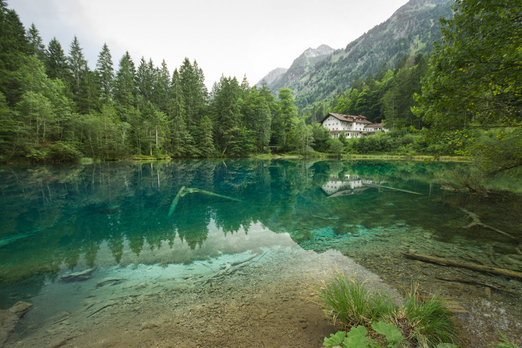 Türkis schimmernder, spiegelnder Christlessee im Allgäu mit Nadelbäumen, Hotel am See und Bergen im Hintergrund