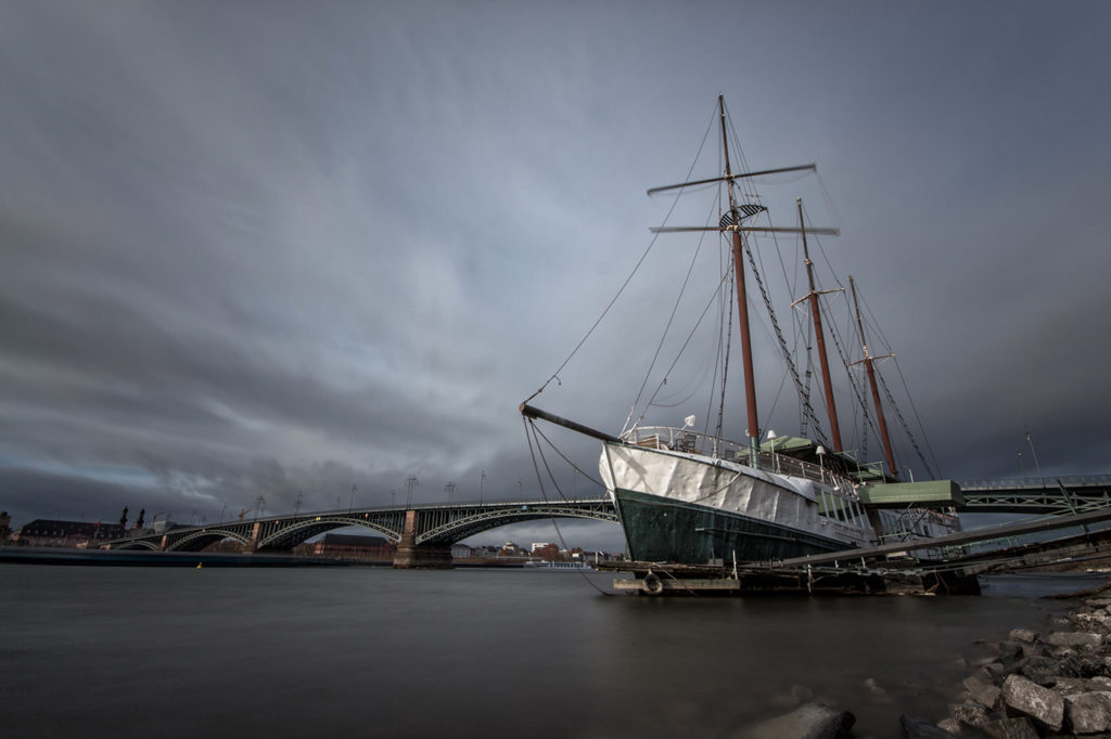 Am Rheinufer liegendes Segelschiff mit Theodor-Heuss-Brücke im Hintergrund